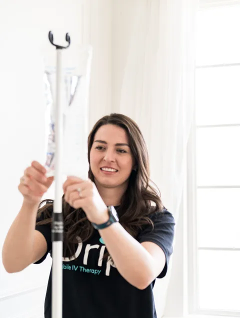 A DrpIV+ nurse in a black Drp tee hanging an IV bag on its pole, smiling
