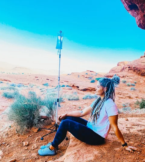 A DrpIV+ client sitting on red desert rock in southern Utah, her IV pole beside her