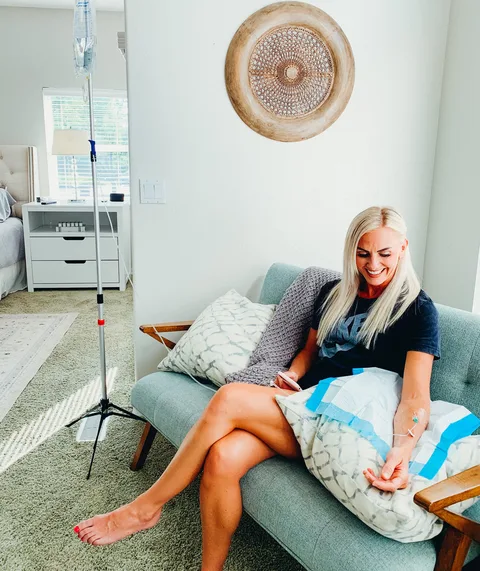 A woman smiling on her couch with a pillow on her lap during her IV