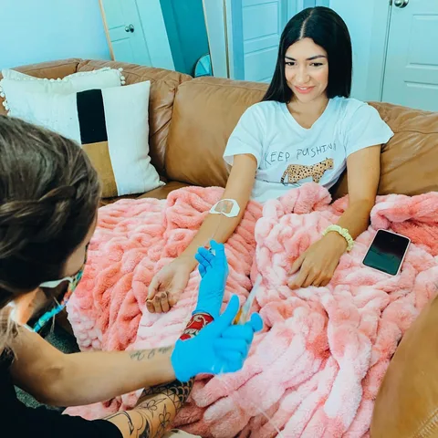 A woman under a pink blanket on her couch while a nurse in blue gloves starts her IV