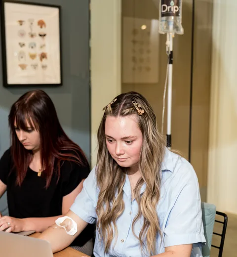 A woman working on her laptop during her IV, a DrpIV+ nurse beside her