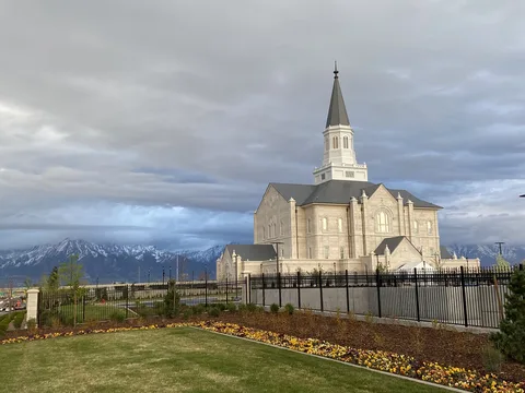 The Taylorsville Utah Temple under spring clouds, with the snowy Wasatch Range behind