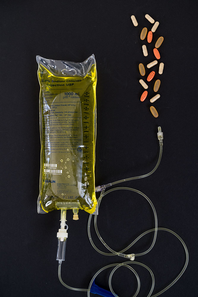 Vitamin Bar IV Bag against a black background. Yellow fluid in the bag and some vitamin pills on the side.