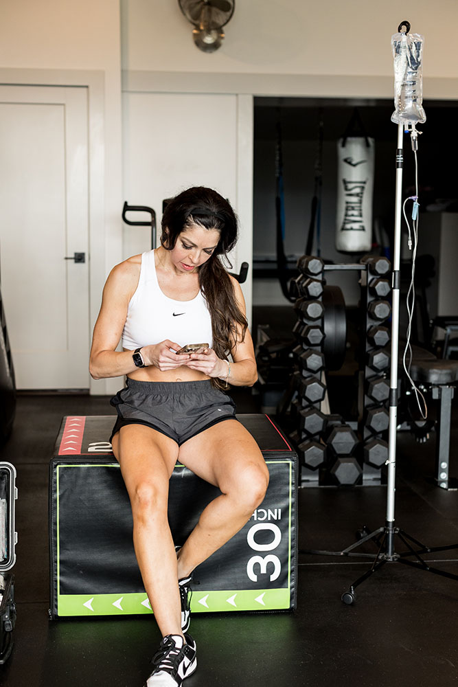 Woman who uses peptides for muscle growth at her home gym sitting on a jump box next to a hanging bag of IV fluid