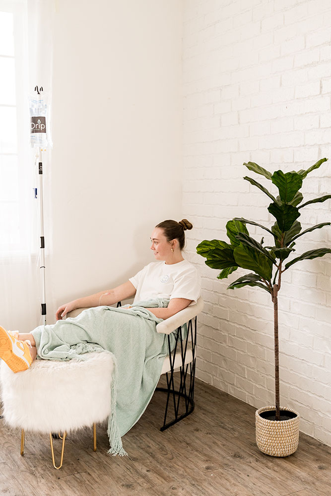 Woman receiving iv therapy for jet lag sitting on a white chair with a plant nearby and the iv fluid bag hung on a pole